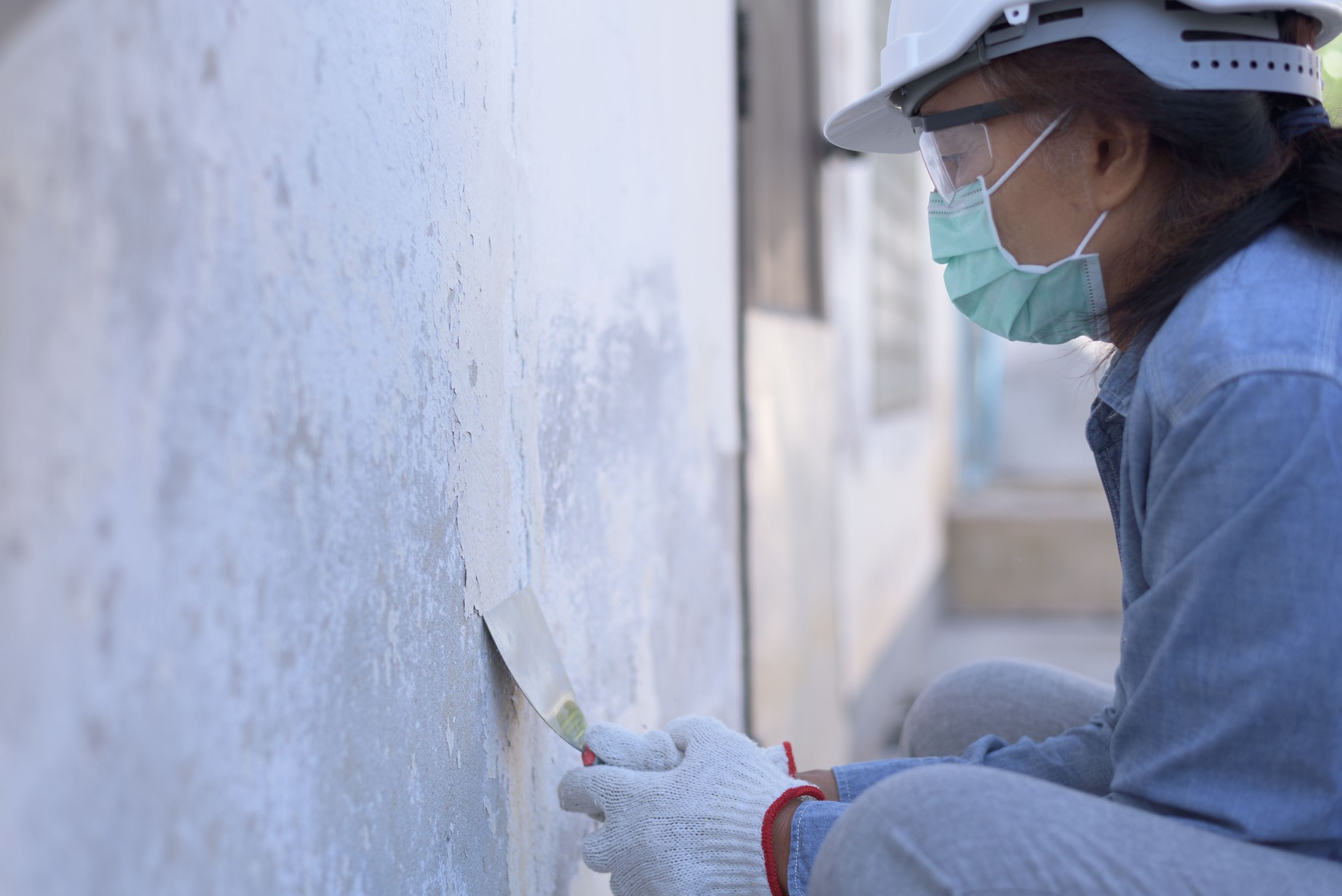 Side view of senior Asian female worker wearing safety hard hat, goggles, gloves and mask, holding trowel, trying to peel off paint film from the wall. Retired lifestyle DIY home renovation concept.